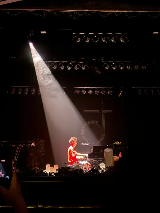 A pianist in a red shirt under a single spotlight on a dark stage