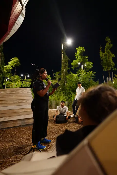 Reading poetry into a microphone outdoors at night, a few people listening